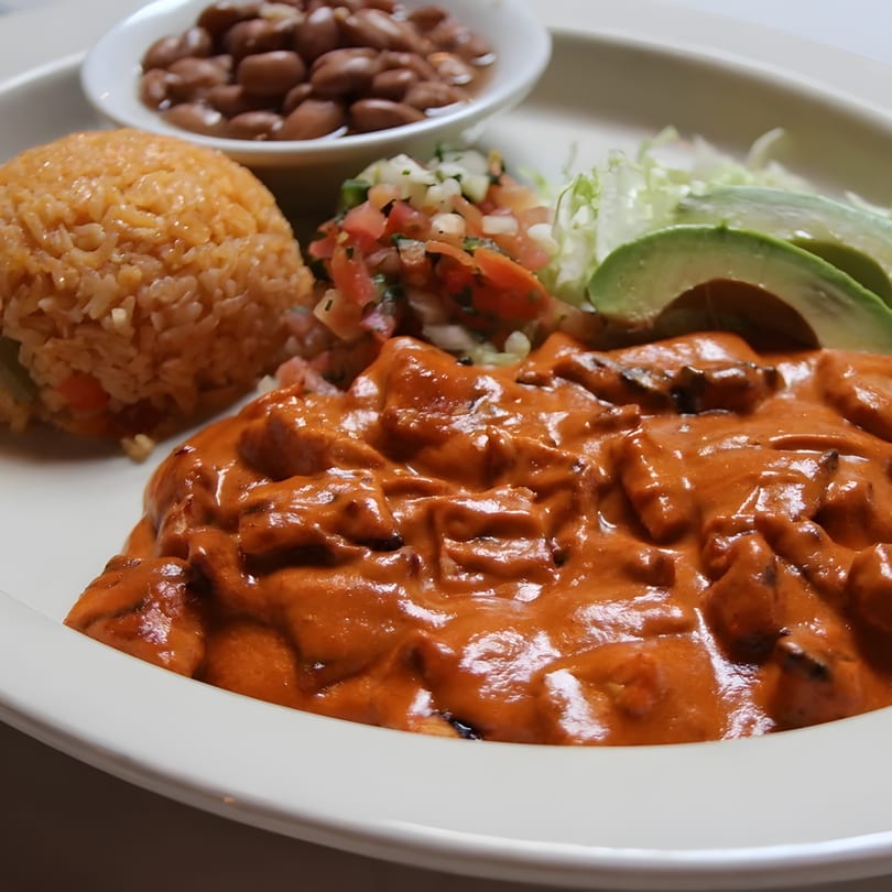 A plate of Mexican food with chicken in red sauce, rice, beans, pico de gallo, and avocado.