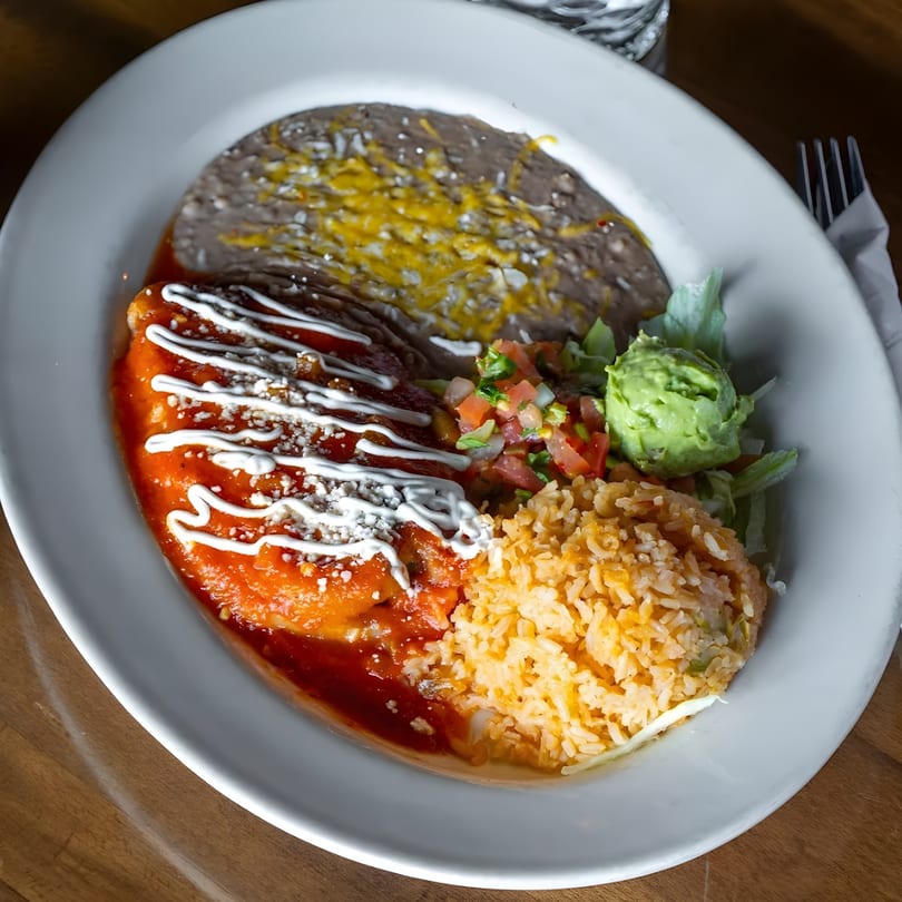 A white plate holds an enchilada with red sauce, rice, refried beans, guacamole, and pico de gallo.