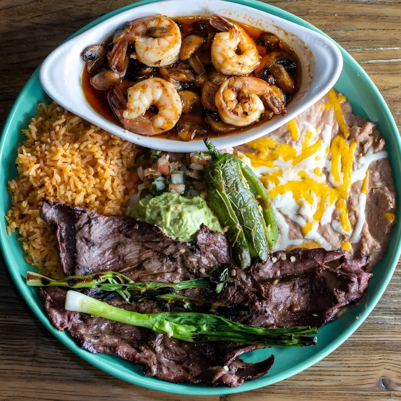 A vibrant Mexican platter with steak, rice, beans, guacamole, pico de gallo, and shrimp in sauce.