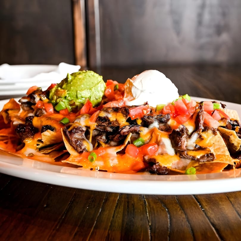 Loaded nachos with melted cheese, steak, diced tomatoes, guacamole, and sour cream on a white plate.